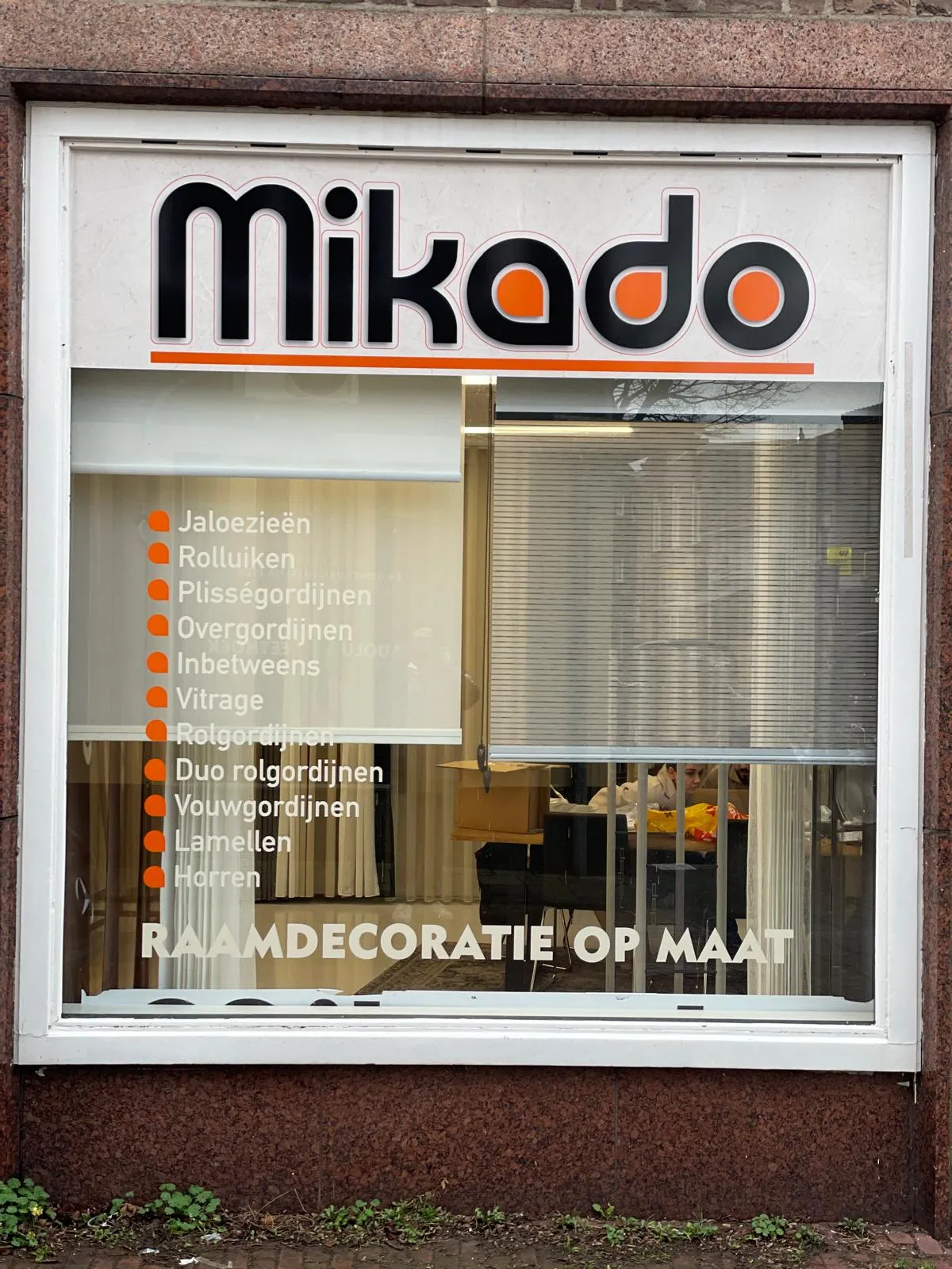 Etalageraam van raamdecoratiewinkel Mikado met het zwart-oranje logo en een productoverzicht op de ruit.