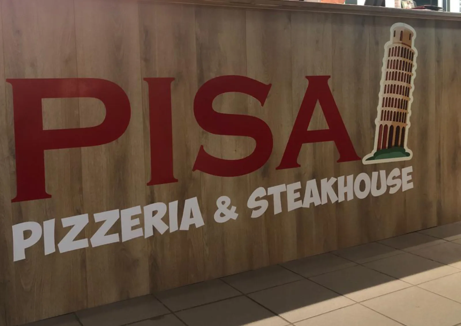 Wandreclame met de rode PISA-belettering, de tekst Pizzeria en Steakhouse en een illustratie van de toren van Pisa op een houten wand.